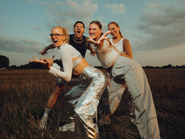 Vier Menschen posieren energisch auf einem Feld unter einem teilweise bewölkten Himmel.