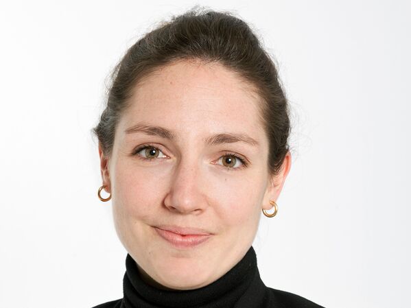 Weiblich gelesene Person mit braunem Haar in einem Dutt, trägt einen schwarzen Rollkragenpullover und einen Blazer, lächelt.