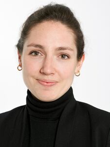 Weiblich gelesene Person mit braunem Haar in einem Dutt, trägt einen schwarzen Rollkragenpullover und einen Blazer, lächelt.