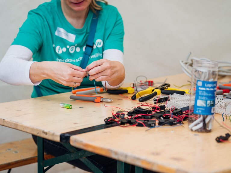 Person, die mit Drähten und Werkzeugen an einem Tisch arbeitet und elektronische Bauteile vorbereitet.