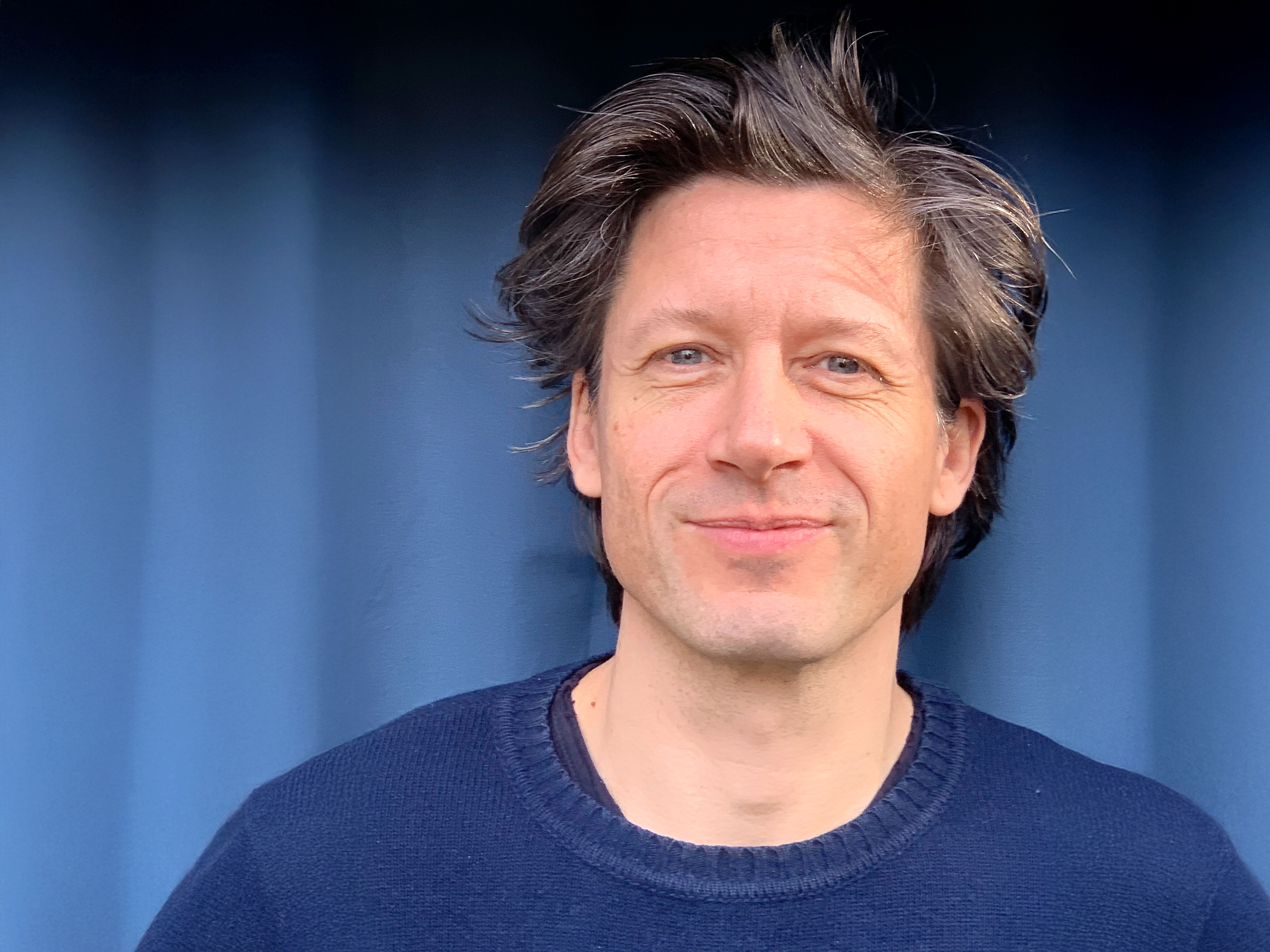 Ein Mann mit mittellangen braunen Haaren und einem blauen Pullover steht vor einem dunklen Hintergrund.