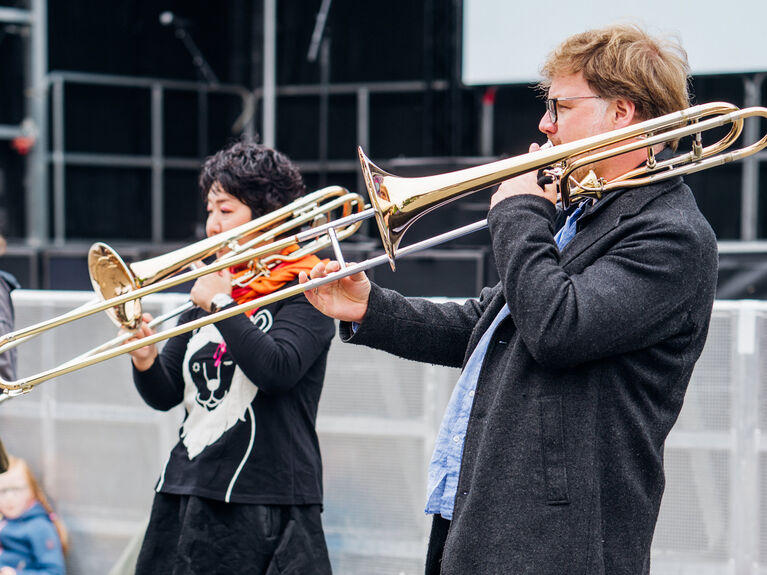 Zwei Personen spielen Posaune im Freien auf einer gepflasterten Fläche in der Nähe einer Bühne.