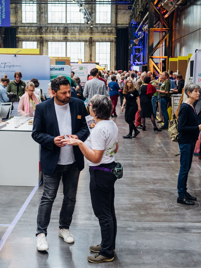 In einer überdachten Ausstellung mit verschiedenen Ständen und Auslagen kommen Menschen zusammen und unterhalten sich.