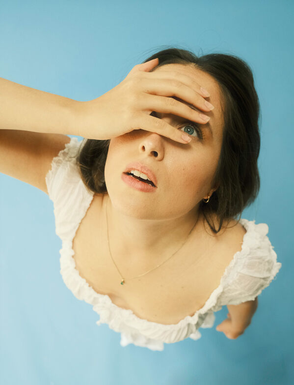 Frau in weißem Oberteil blickt mit einer Hand auf ihre Stirn vor blauem Hintergrund.