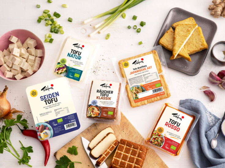Verschiedene Tofuprodukte mit Gemüse und Kräutern auf einer weißen Tischplatte.