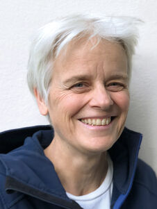 Eine Person mit kurzen weißen Haaren lächelt, sie trägt eine dunkle Jacke, vor einer hellen Wand.