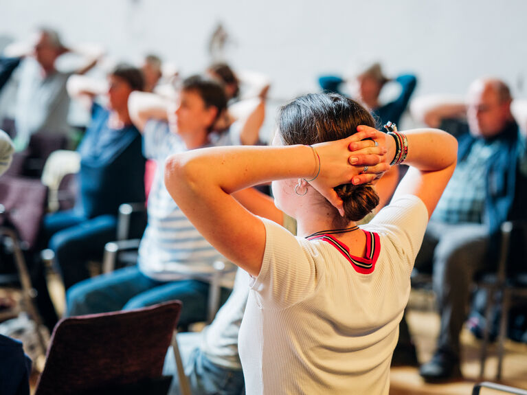 Menschen, die in einer Gruppe auf Stühlen sitzen und ihre Hände hinter dem Kopf verschränken.
