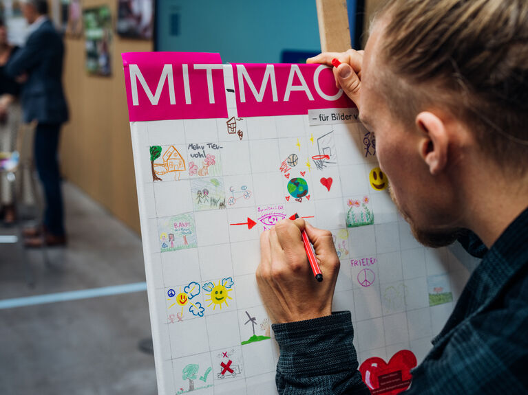Eine Person zeichnet auf eine bunte Plakatwand mit der Aufschrift MITMACH in einem öffentlichen Raum.