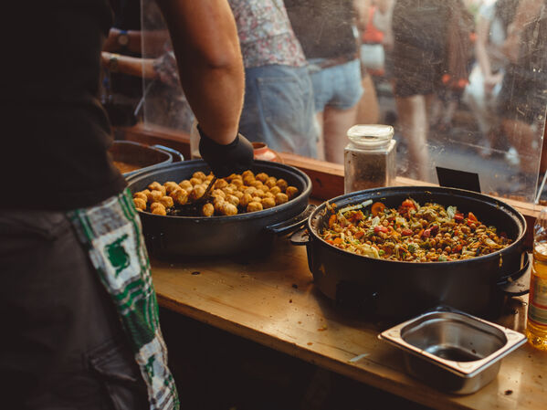Eine Person serviert Essen aus großen Pfannen an einem Marktstand im Freien.