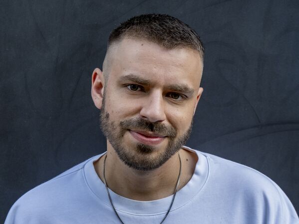 Männlich gelesene Person mit kurzen Haaren und Bart, trägt ein weißes Sweatshirt und eine Halskette, blickt in die Kamera.