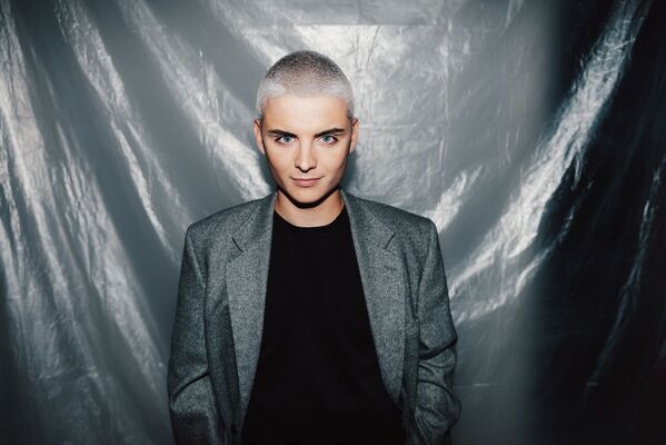 Eine Person mit kurzen Haaren in einem grauen Blazer steht vor einem Plastikhintergrund.