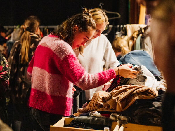 Zwei Personen sehen sich die Kleidung auf einem Tisch in einer belebten Markthalle oder bei einem Verkauf an.