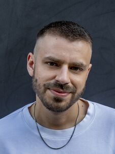 Männlich gelesene Person mit kurzen Haaren und Bart, trägt ein weißes Sweatshirt und eine Halskette, blickt in die Kamera.